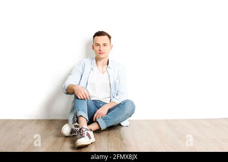 Porträt eines glücklichen jungen Millennials mit strahlenden Lächeln in einem weißen Hemd ohne Knöpfe sitzt auf einem Holzboden, isoliert. Selbstbewusster lässiger Hippie-Look Stockfoto