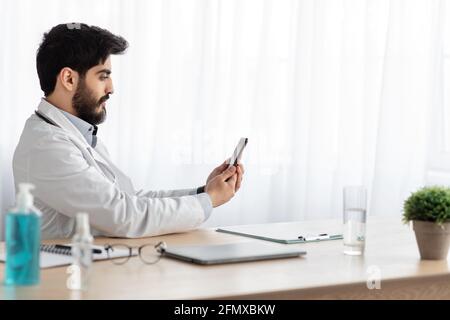 Ruhiger gutaussehender junger arabischer bärtiger Arzt im weißen Mantel Betrachtet die Webcam des Smartphones Stockfoto