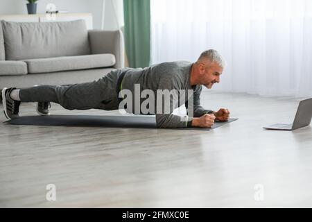 Athletischer bärtiger älterer Mann, der zu Hause mit einem Laptop Übungen macht Stockfoto