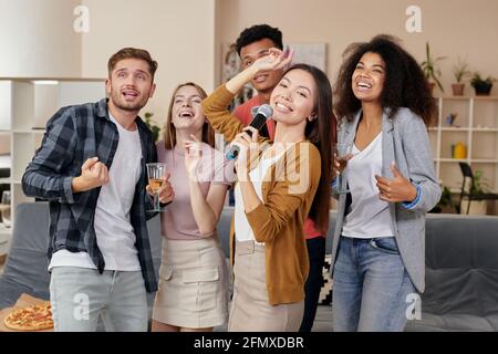 Party-Spaß. Junge asiatische Mädchen hält Mikrofon und singen, während Karaoke mit besten Freunden zu Hause spielen. Gruppe von Menschen trinken, Spaß haben Stockfoto