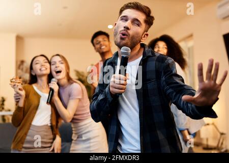 Singe gern. Enthusiastisch schöner junger Mann, der mit Mikrofon singt, während er mit Freunden zu Hause Karaoke spielt und in der modernen Wohnung steht Stockfoto