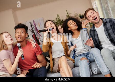 Singen Sie, wie Sie können. Eine enthusiastische Gruppe von Freunden singt mit Mikrofon, während sie zu Hause Karaoke spielen und zu Hause auf der Couch sitzen Stockfoto