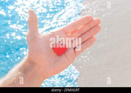 In der Hand eines Mannes auf der Handfläche eines kleinen roten Herzens Nahaufnahme in Form eines Schlosses. Ein Zeichen oder Symbol der Liebe gegen die Küste. Stockfoto