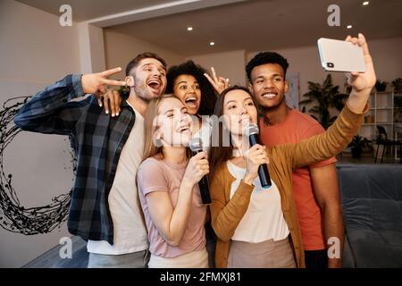 Selfie-Zeit. Eine Gruppe junger, fröhlicher, multikultureller Freunde, die mit dem Smartphone ein Selfie machen, während sie Zeit miteinander verbringen und zu Hause Karaoke spielen Stockfoto