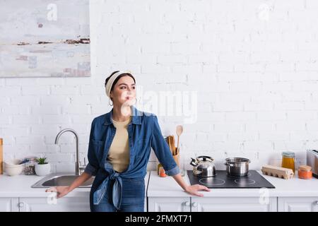 Lächelnde Hausfrau im Denim-Hemd, das neben der Küchenarbeitsfläche schleift Stockfoto