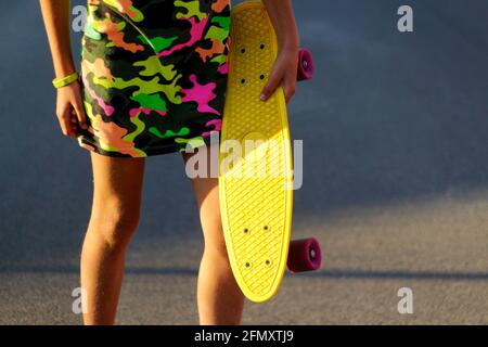 Defokussieren Mädchen mit gelben Penny Board. Vorderansicht. Sommer in leuchtenden Farben. Kinderhände halten tagsüber ein kurzes Cruiser-Pennyboard im Freien. Urbane Mode Stockfoto