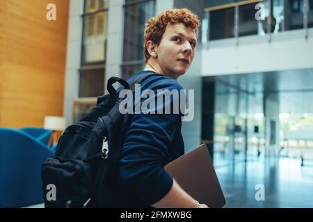Student mit Tasche und Laptop auf dem Campus. Junger Mann, der sich über die Schulter dreht und auf dem College-Campus läuft. Stockfoto