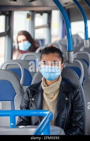 Eine Frau, die in einem Bus mit einer Gesichtsmaske unterwegs ist, und eine andere Frau, die weiter hinten im Bus sitzt. Stockfoto