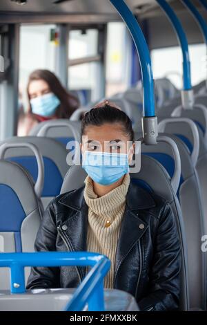 Eine Frau, die in einem Bus mit einer Gesichtsmaske unterwegs ist, und eine andere Frau, die weiter hinten im Bus sitzt. Stockfoto