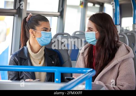 Zwei Frauen in ihren zwanzig Jahren tragen Gesichtsmasken und unterhalten sich mit ihnen Jeder in einem Bus Stockfoto