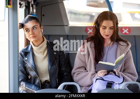 Zwei Frauen mit Schutzmasken sitzen auf einem Bus Einer schaut nebeneinander aus dem Fenster und der andere Lesen Stockfoto