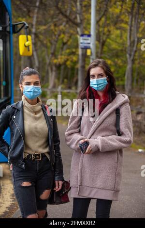 Zwei junge Frauen mit Gesichtsbezügen warten vor einem Bus an einer Bushaltestelle. Stockfoto