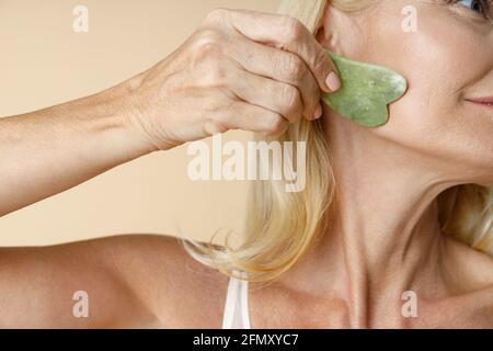 Nahaufnahme einer reifen blonden Frau mit perfektem Leuchten Haut mit Jade Gua Sha Massagewerkzeug für die Hautpflege Und Schönheitsbehandlungen isoliert über Beige Stockfoto