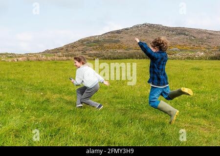 Aufgeregte, energische Kinder, die durch ein Feld mit Zennor Hill im Hintergrund in Cornwall laufen. Stockfoto