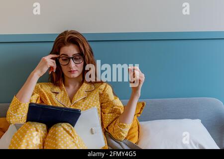 Eine junge Frau im bequemen gelben Pyjama arbeitet, während sie im Bett liegt. Geschäftsfrau pflegt eine soziale Distanzierung arbeitet aus der Ferne. Neues normales Konzept Stockfoto