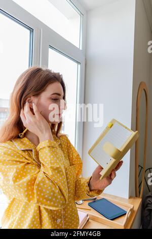 Eine junge Frau im bequemen gelben Pyjama sitzt auf der Fensterbank im Schlafzimmer. Geschäftsfrau unterstützt soziale Distanzierung, arbeitet aus der Ferne. Neues normales Konzept Stockfoto