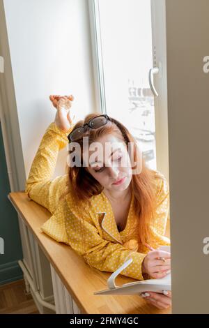 Eine junge Frau im bequemen gelben Pyjama arbeitet und liegt auf der Fensterbank im Schlafzimmer. Geschäftsfrau unterstützt soziale Distanzierung, arbeitet aus der Ferne. Neues normales Konzept Stockfoto