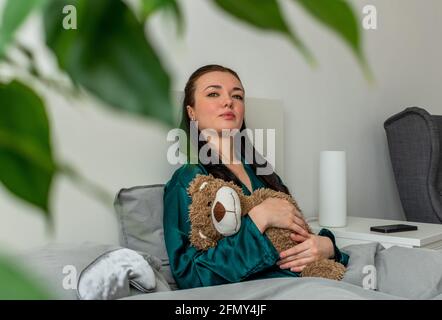 Eine junge Frau im Seidenpyjama sitzt auf dem Bett und umarmt einen Teddybären. Zurück zu den Wurzeln. Wellness-Konzept. Stockfoto