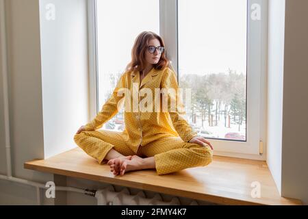 Junge Geschäftsfrau meditiert auf der Fensterbank im Schlafzimmer liegend. Mädchen hört ruhige Musik. Home Komfort Zone und Wellness-Konzept. Stockfoto