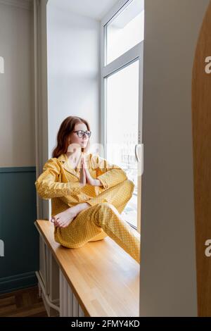 Junge Geschäftsfrau meditiert auf der Fensterbank im Schlafzimmer liegend. Mädchen hört ruhige Musik. Home Komfort Zone und Wellness-Konzept. Stockfoto