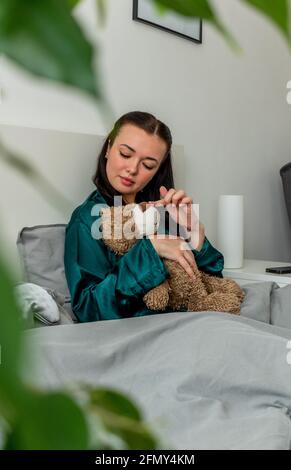 Eine junge Frau im Seidenpyjama sitzt auf dem Bett und umarmt einen Teddybären. Zurück zu den Wurzeln. Wellness-Konzept. Stockfoto