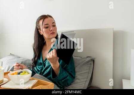 Die junge Frau im Seiden-Pyjama frühstückt gesund und liest im Schlafzimmer auf dem Bett. Stockfoto