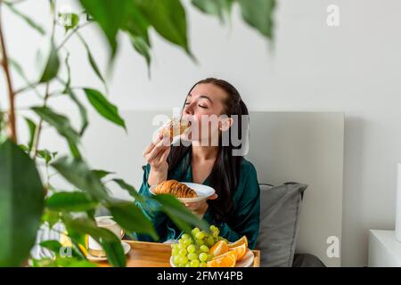 Die junge Frau im Seiden-Pyjama hat ein gesundes Frühstück, während sie im Schlafzimmer auf dem Bett liegt. Konzept für das Wohlbefinden zu Hause. Emotionale Gesundheit einer jungen Frau Stockfoto