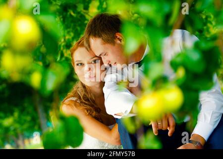 Wunderschöne Brautjungfern umarmen und lächeln in einem grünen Garten mit Bäumen. Rothaarige hübsche Frau in einem weißen Kleid, ein Mann im weißen Hemd, eine schwarze Fliege. Stockfoto
