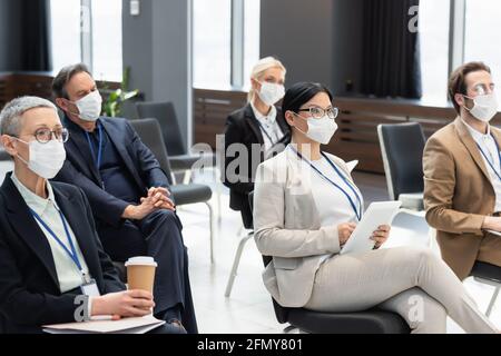 Interracial Geschäftsleute in medizinischen Masken sitzen auf Seminar mit Coffee to go und digitales Tablet Stockfoto