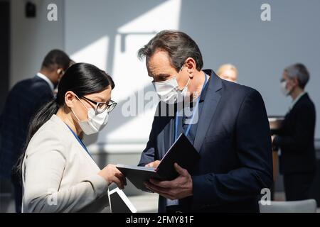 Reifer Geschäftsmann in medizinischer Maske zeigt Notizbuch zu asiatischen Kollegen Stockfoto