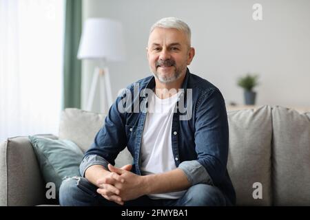Schöner grauhaariger älterer Mann, der im Wohnzimmer auf der Couch posiert Stockfoto