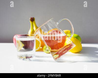 Kanne mit reinem Wasser, das auf dem Rand steht, Wasser in das Glas gießen. Set von frischen Früchten auf dem Hintergrund. Stockfoto