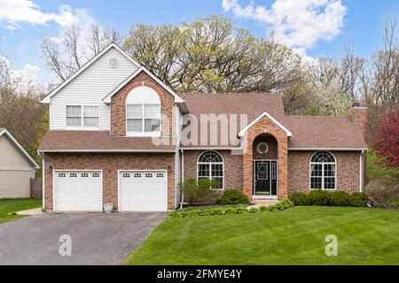 Ein vorstädtisches, rotes Backsteinhaus und weißes Vinyl-Haus mit einer Garage für zwei Autos, einem Gipfel über der Eingangstür und einer gepflegten Landschaft. Stockfoto