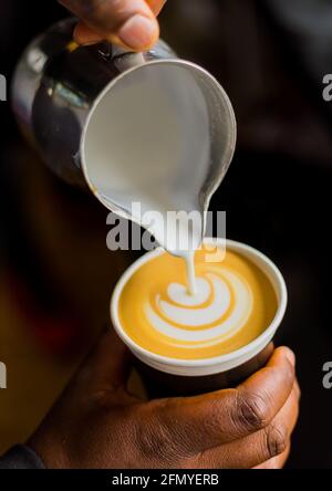Vertikale Aufnahme eines afrikanischen Kaffee-Barista, der ein Blatt ausgießt Formen Sie die Form mit Milchschaum in einer Tasse zum Mitnehmen Stockfoto