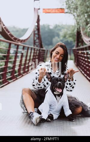 Eine schöne junge Frau in einem flauschigen Rock und einer gepunkteten Bluse sitzt auf der Brücke mit ihrem Staffordshire Bull Terrier Hund, der lustig lächelt. Softselect Stockfoto