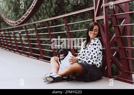 Eine schöne junge Frau in einem flauschigen Rock und einer gepunkteten Bluse sitzt mit ihrem Staffordshire Bull Terrier Hund auf der Brücke. Weicher, selektiver Fokus. Kunst Stockfoto