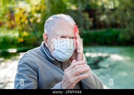 Kranker alter grauhaariger Mann sitzt in einer medizinischen Maske. Männer haben Kopfschmerzen. Konzept von Kopfschmerzen. Stockfoto