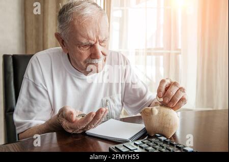 Der alte Mann in der Pandemie zählt Pennys aus dem Pensionsfonds. Ein älterer Mann, der über die Sperrung besorgt ist, steckt Geld in eine Sparschwein. Rentner rechnen damit Stockfoto