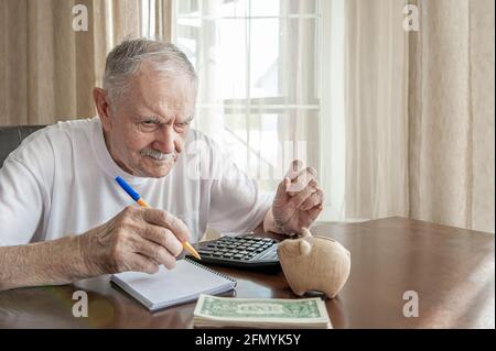 Der alte Mann mit Bleistift zählt Pfennige aus der Pensionskasse, die an einem Tisch im Zimmer sitzt. Besorgter älterer Mann steckt Geld in eine Sparschwein. Rentner c Stockfoto