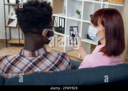 Multirassisches Paar in Gesichtsmaske, das auf der Couch sitzt und Videogespräche mit muslimischen Freunden führt. Diverse Freunde, die moderne Gadgets für die Fernkommunikation während einer Pandemie verwenden. Stockfoto