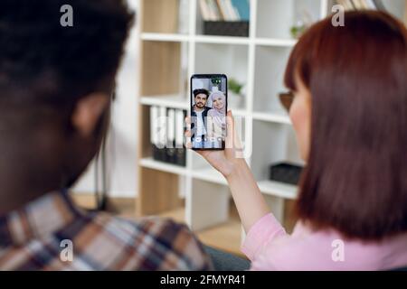 Betrachten Sie von der Schulter eines multirassischen Paares, das mit einem modernen Smartphone in den Händen auf der Couch sitzt und mit muslimischen Freunden per Videoanruf spricht. Konzept von Mensch und Technik. Stockfoto