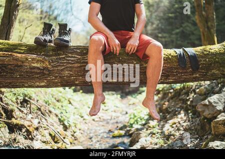 Mann, der auf dem gefallenen Baumstamm über dem Bergwaldbach sitzt, während er auf Socken wartet, die trocknen und Trekkingstiefel. Aktive Menschen reisen Stockfoto