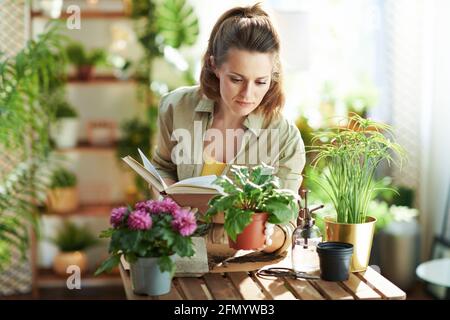 Green Home. Moderne 40 Jahre alte Hausfrau in weißen Gummihandschuhen mit Topfpflanze und Buch zu Hause in modernen sonnigen Tag. Stockfoto