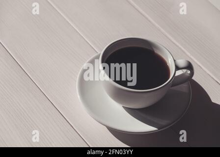 Schwarzer Kaffee in weißer Tasse mit Untertasse auf hellem Holztisch, Schatten, Kopierraum Stockfoto
