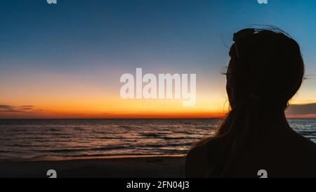 Frau beobachtet den Sonnenuntergang über dem Meeresstrand in Silhouette von hinten Stockfoto