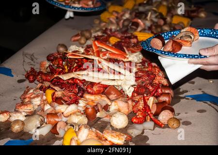Hand halten Pappteller mit ausgewählten Stücken von Lebensmitteln aus Großer Stapel von Meeresfrüchten und Würstchen sowie Kartoffeln und Pilzen Auf einem papierbedeckten Tisch gestapelt Stockfoto