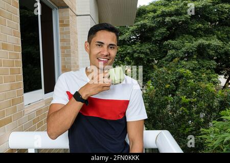 Lächelnder junger Brasilianer, der eine Tasse Kaffee auf dem Balkon des Gebäudes hält. Stockfoto