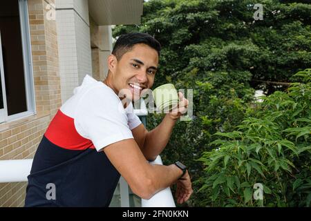 Lächelnder junger Brasilianer, der eine Tasse Kaffee in der Hand hält und sich auf die Balustrade des Balkons des Gebäudes lehnt. Stockfoto