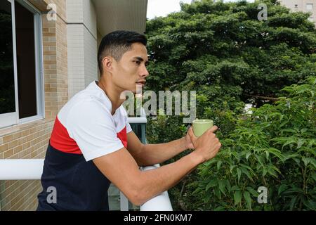 Junger Brasilianer, der eine Tasse Kaffee in der Hand hält, blickt nach vorne und lehnt sich auf die Balustrade des Balkons des Gebäudes. Stockfoto