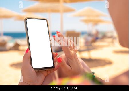 Frau benutzt am Strand ein Mockup-Handy. Frau hält am Strand ein Handy mit leerem Bildschirm. Nahaufnahme der weiblichen Hände, die Reisende benutzen Stockfoto